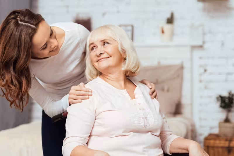 A young woman stands beside an older woman seated in a wheelchair, gently placing her hands on the older womans shoulders. They are smiling at each other in a cozy, well-lit room.