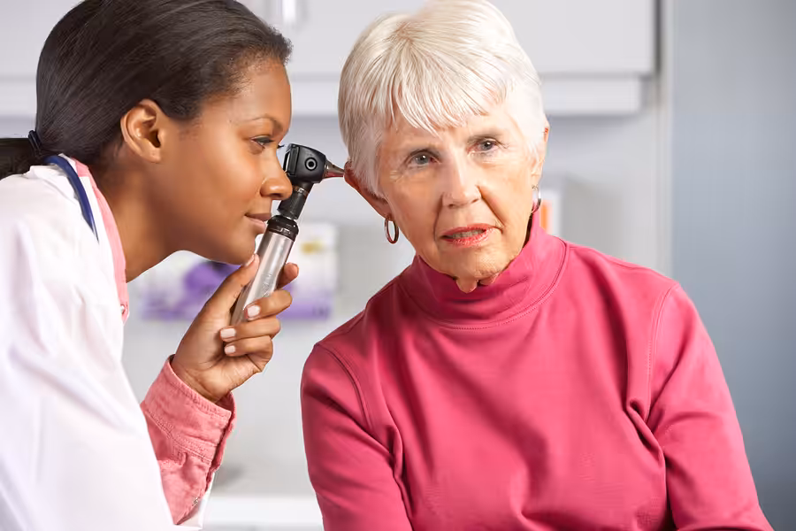 A female doctor examines the ear of an older woman with a medical otoscope in a clinical setting. The patient, wearing a pink turtleneck, looks slightly concerned.