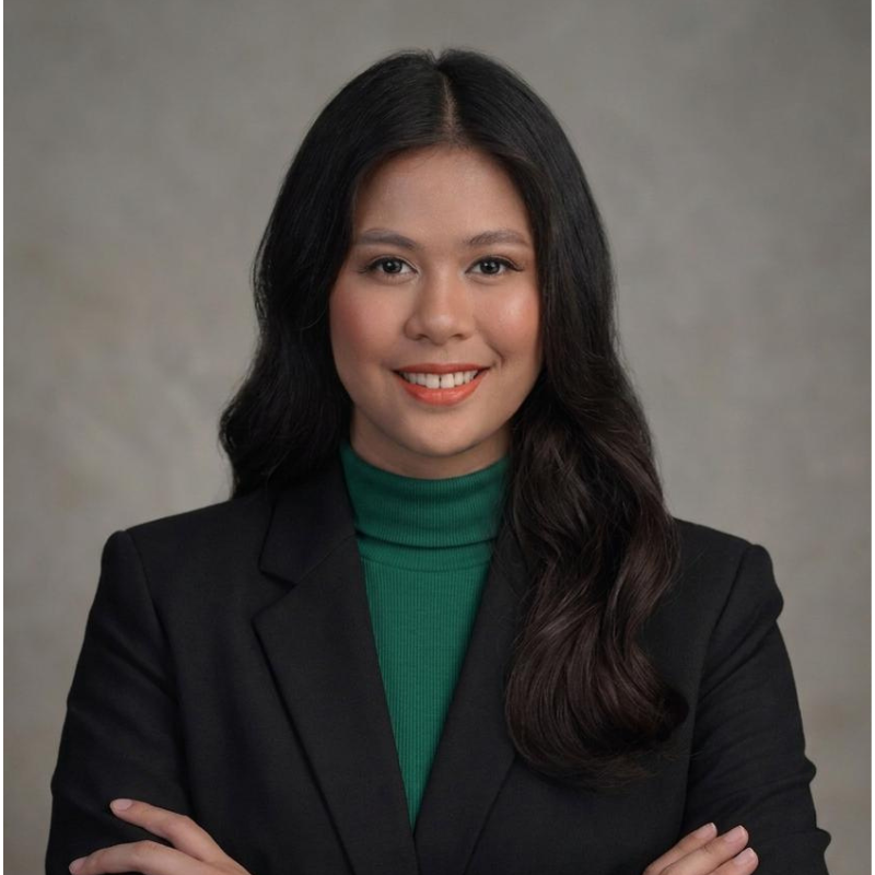 A woman with long dark hair, wearing a green turtleneck and black blazer, smiles with arms crossed in front of a plain, neutral background.