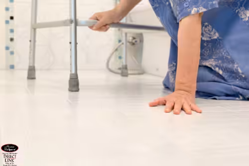 An elderly person wearing a blue robe is on the bathroom floor, reaching toward a walker for support, appearing to have fallen and trying to get up.