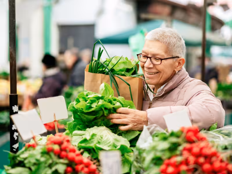 An older adult with short gray hair smiles while shopping at an outdoor market, holding a paper bag filled with leafy greens and green onions, surrounded by fresh produce including lettuce and radishes.