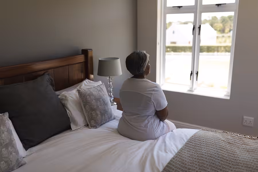 An older woman sits on the edge of a neatly made bed, looking out of a bright window in a calm, softly lit bedroom decorated in neutral tones.