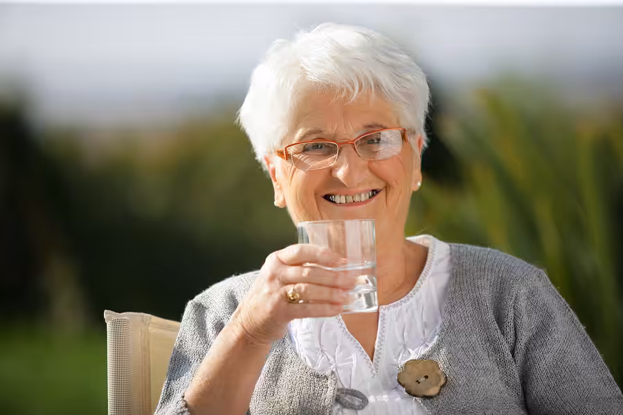 Smiling elderly woman with short white hair and glasses, sitting outdoors, holding a glass of water. She is wearing a gray cardigan over a white blouse and appears relaxed and happy. Greenery is blurred in the background.