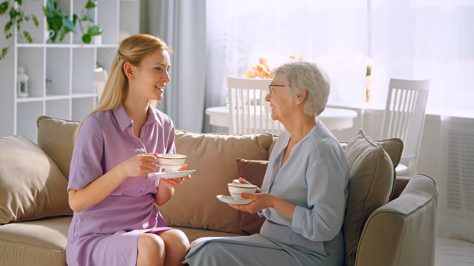 A young woman and an older woman sit on a sofa, smiling and holding teacups, enjoying a conversation in a bright, cozy living room.