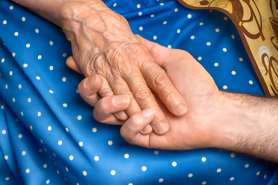 A younger person gently holds the wrinkled hand of an older person, both resting on a blue fabric with white polka dots. The image conveys care, support, and compassion between generations.