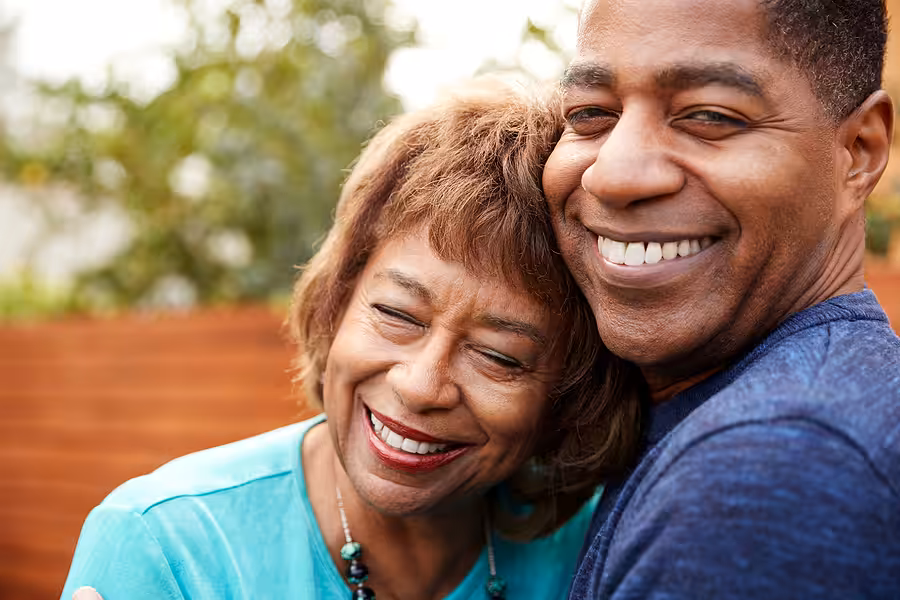 A smiling older woman and a smiling middle-aged man hug outdoors. The woman has short brown hair and wears a turquoise top, while the man wears a blue shirt. Both look happy and content.