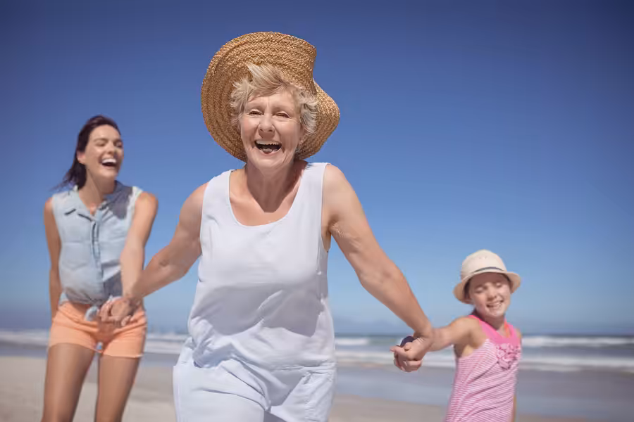 Three women of different ages, including a senior, an adult, and a child, are smiling and holding hands while walking on a sunny beach. They all wear summer clothes and hats, enjoying a joyful day together.
