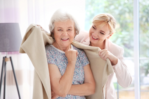 A smiling elderly woman stands indoors while a younger woman gently drapes a beige blanket over her shoulders. They both appear happy and are in a bright room with a lamp and window in the background.