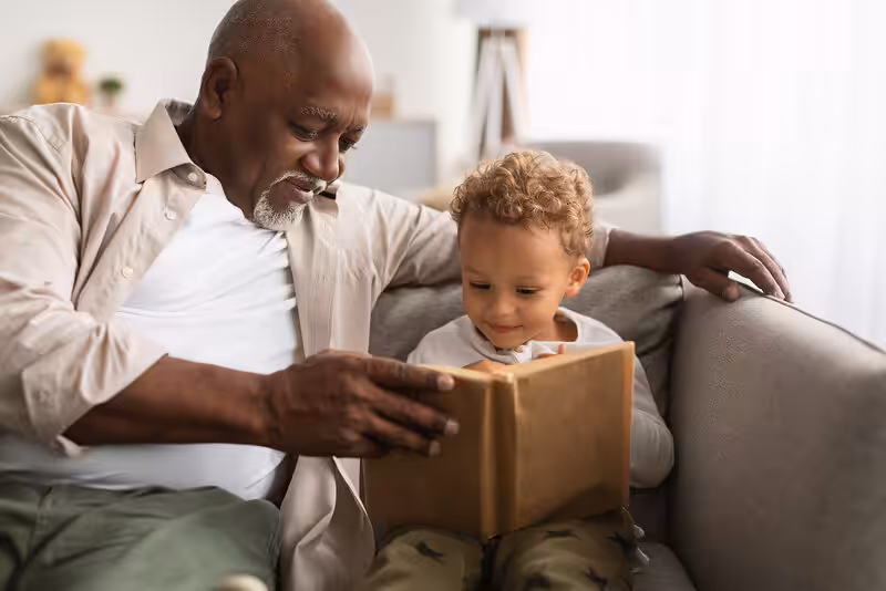 An older man and a young boy sit together on a couch, smiling and reading a book. The man points at the book as the boy looks on attentively. They appear to be enjoying their time together.