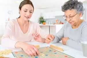 Two women, one younger and one older, sit at a table playing a board game together in a bright, modern kitchen. The younger woman is placing a tile on the board, while the older woman watches.