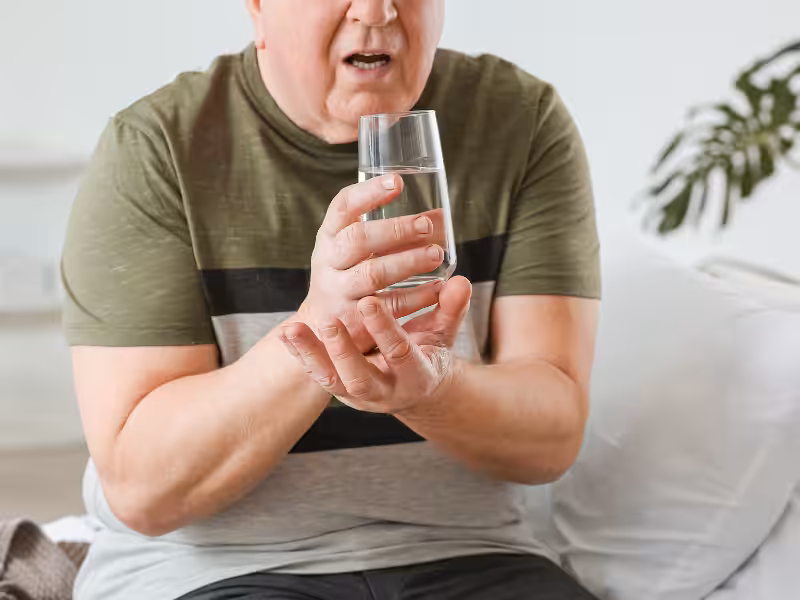 An older adult sits on a couch, holding a glass of water with both hands. His hands are shaking, and he appears to struggle with the task. A pillow and plant are visible in the background.