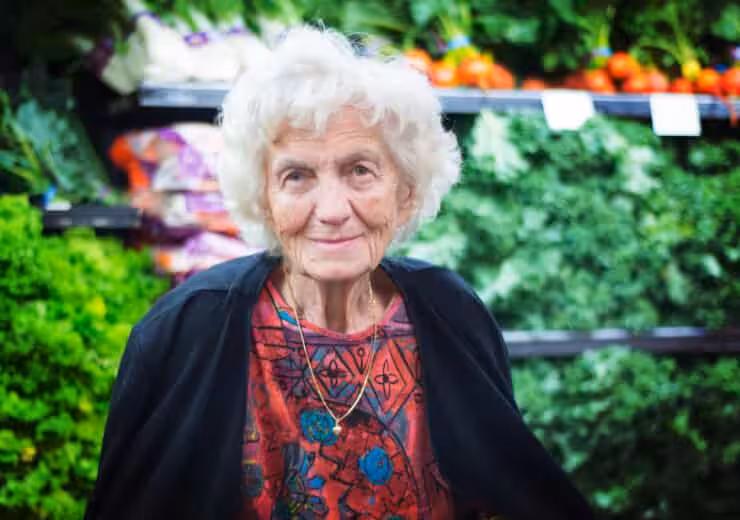 An elderly woman with white hair, wearing a colorful red patterned top and a dark cardigan, stands in front of a display of fresh leafy greens and vegetables at a grocery store.