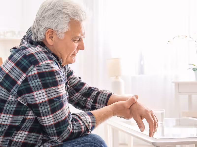 An older man in a plaid shirt sits on a couch, holding his wrist with a pained expression. A glass of water sits on a nearby table in a softly lit, cozy room.