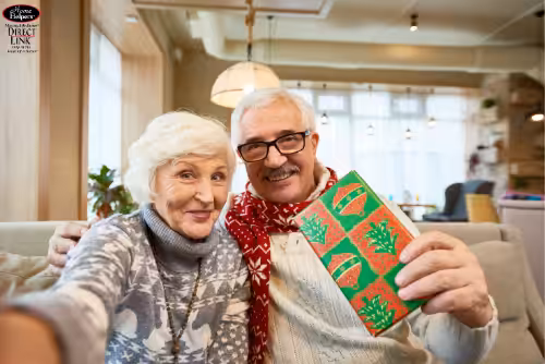 An elderly couple sits on a couch, smiling at the camera. The woman wears a gray sweater, while the man wears glasses, a cream sweater, and a red scarf. He is holding a green and red wrapped gift. The room looks cozy and bright.
