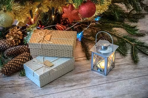 Two wrapped gifts sit under a decorated Christmas tree with pinecones and ornaments, next to a lit lantern on a wooden floor.
