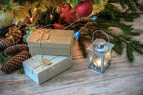 Two wrapped gifts sit under a decorated Christmas tree with pinecones and ornaments, next to a lit lantern on a wooden floor.