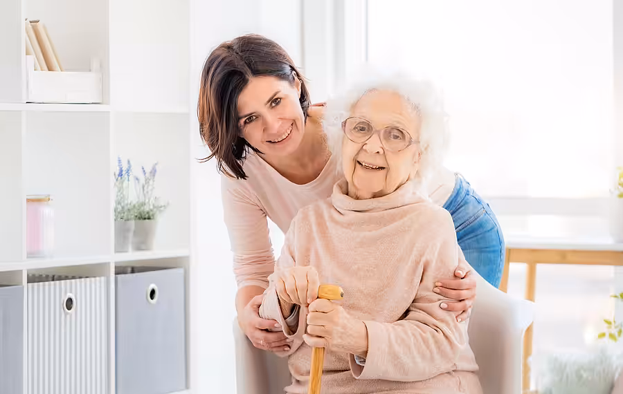 A smiling young woman hugs an elderly woman with white hair and glasses, who is sitting and holding a wooden cane. They are indoors in a bright, cozy room with shelves and plants in the background.