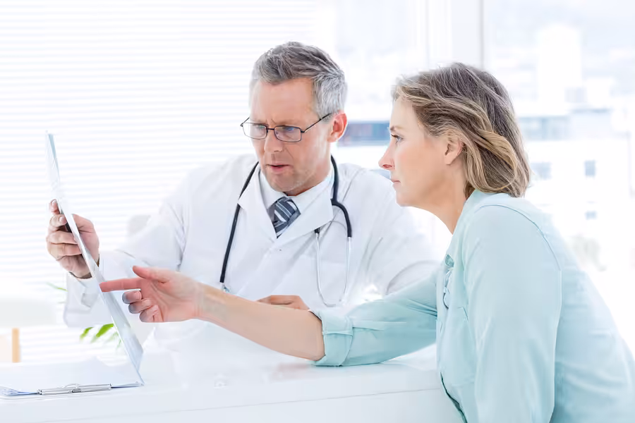 A doctor wearing a white coat and stethoscope discusses medical results with a concerned woman, both looking at a clipboard as the woman points at the document in a bright office setting.