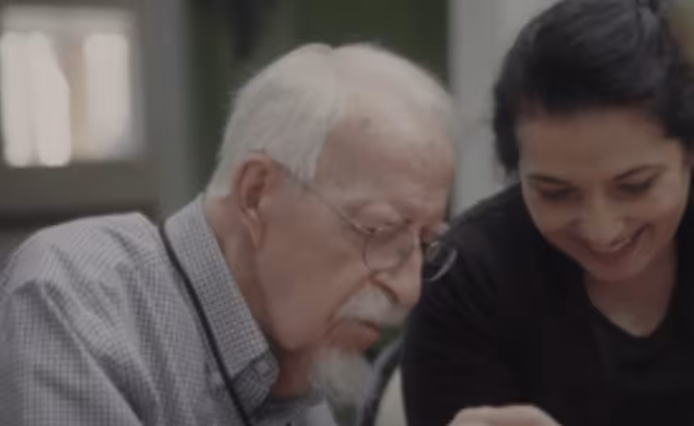 An elderly man with white hair and glasses sits beside a smiling younger woman with dark hair, both engaged in an activity together at a table.