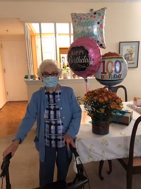 An elderly woman wearing a mask and cardigan stands beside a walker in a decorated room. Behind her are birthday balloons, including one that says “100 Birthday,” and a table with flowers and gifts.
