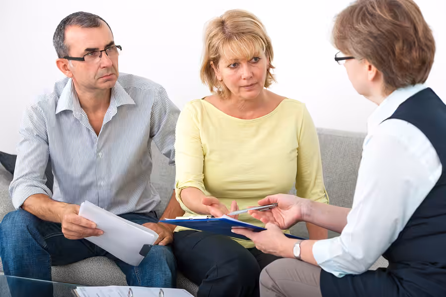 A middle-aged couple sits on a couch, attentively listening to a professional woman who is showing them documents. The couple looks concerned and engaged in conversation.