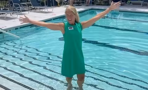 A woman in a green dress stands in a swimming pool with her arms outstretched, smiling. Lounge chairs and a fence are visible in the background.