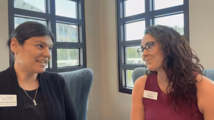 Two women sit facing each other in armchairs by large windows, having a conversation. Both wear name tags and are smiling, suggesting a friendly, professional interaction in a bright office setting.