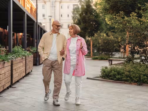 An older couple smiles and walks arm in arm through a city park, dressed casually in light-colored outfits. Lush greenery and modern buildings surround them on a sunny day.