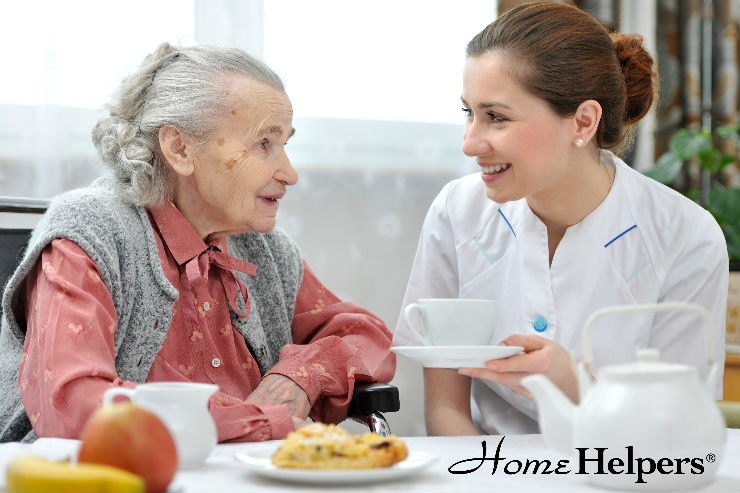 A smiling caregiver in a white uniform serves tea to an elderly woman with gray hair, who is also smiling. They sit at a table with food and a teapot. The image features the Home Helpers logo.
