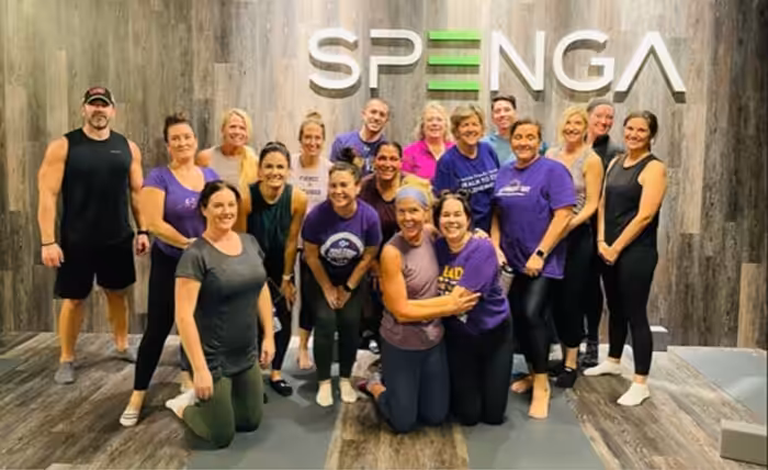 A group of people in workout attire pose and smile together inside a fitness studio with a SPENGA sign on the wall behind them. The group includes men and women of various ages.