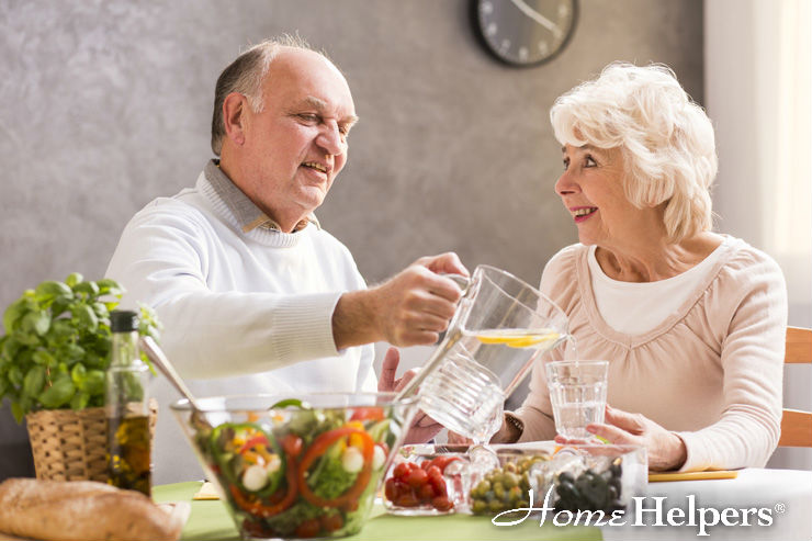 An older man pours water into a glass for an older woman at a dining table with salad, vegetables, and bread. Both are smiling and enjoying each others company. The image is branded with the Home Helpers logo.