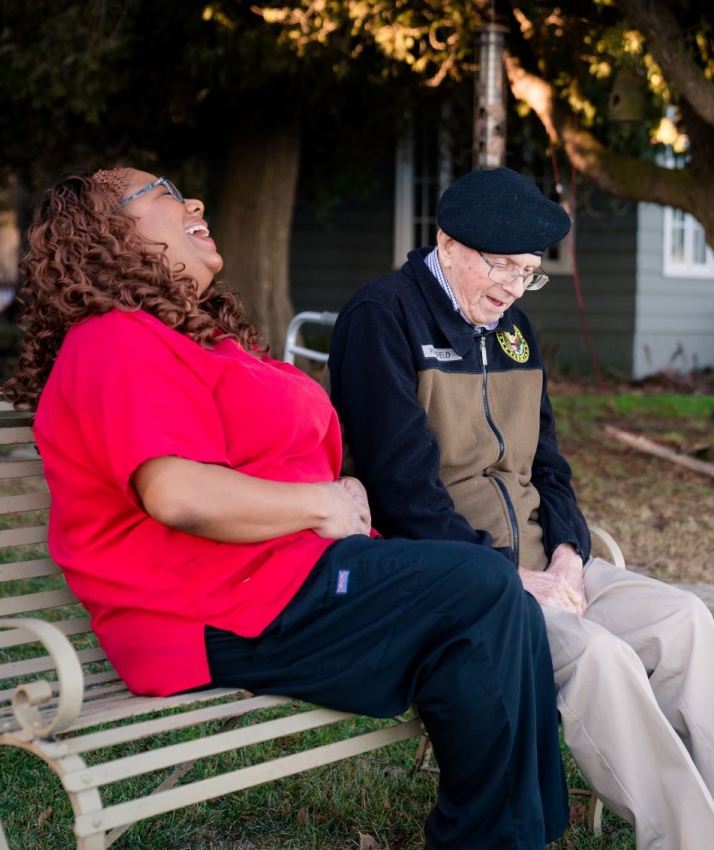 A woman in a red shirt and a man in a black beret sit on an outdoor bench, both smiling and laughing, enjoying each others company on a sunny day.