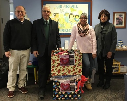 Four adults stand indoors behind two large boxes decorated with colorful, festive wrapping paper and heart-shaped tags. A children’s drawing is framed on the wall behind them. The setting looks cheerful and welcoming.