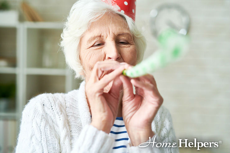 An elderly woman wearing a red party hat and white sweater blows a party horn, smiling. The image has the Home Helpers logo in the bottom right corner.