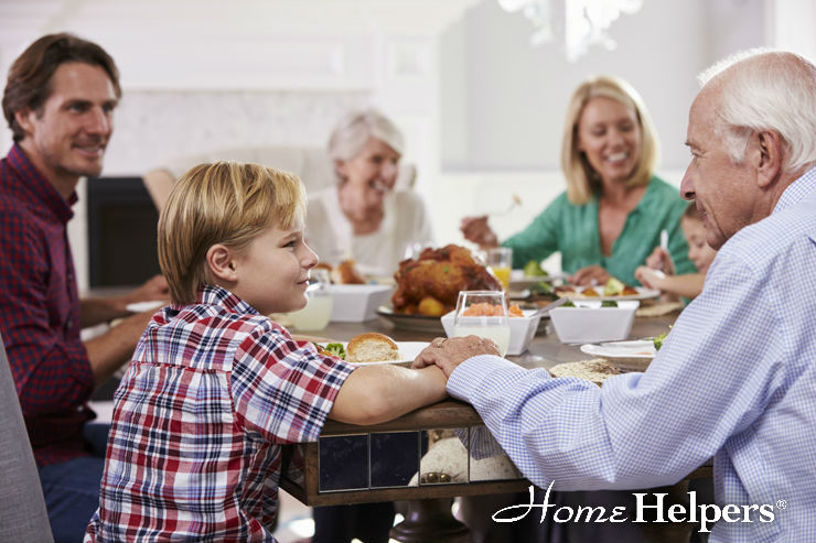 A multigenerational family sits around a dining table sharing a meal, smiling and talking. Food dishes are on the table and an older man holds hands with a young boy. The Home Helpers logo appears in the bottom right corner.
