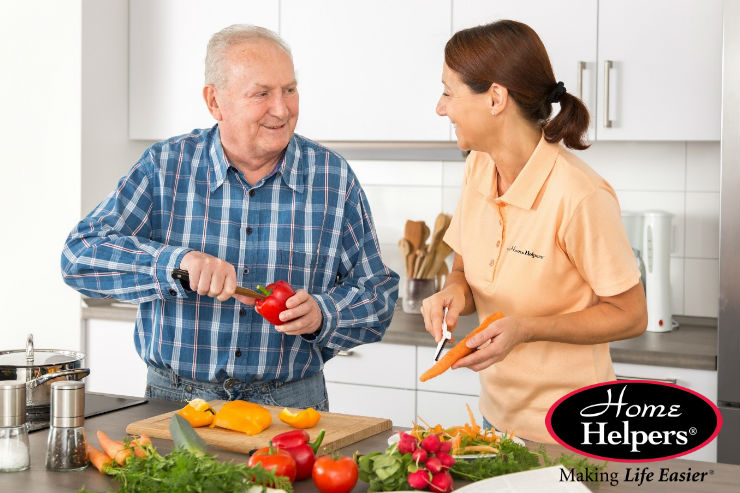 An older man and a woman smile while preparing fresh vegetables together in a bright kitchen. The woman wears a polo shirt with a Home Helpers logo. The Home Helpers logo and the phrase Making Life Easier appear in the corner.