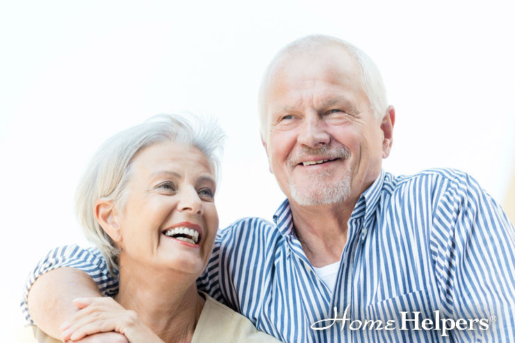 Smiling older couple standing close together, with the man wearing a striped shirt and the woman in a light top. Both appear happy and relaxed. Home Helpers logo is visible in the bottom right corner.