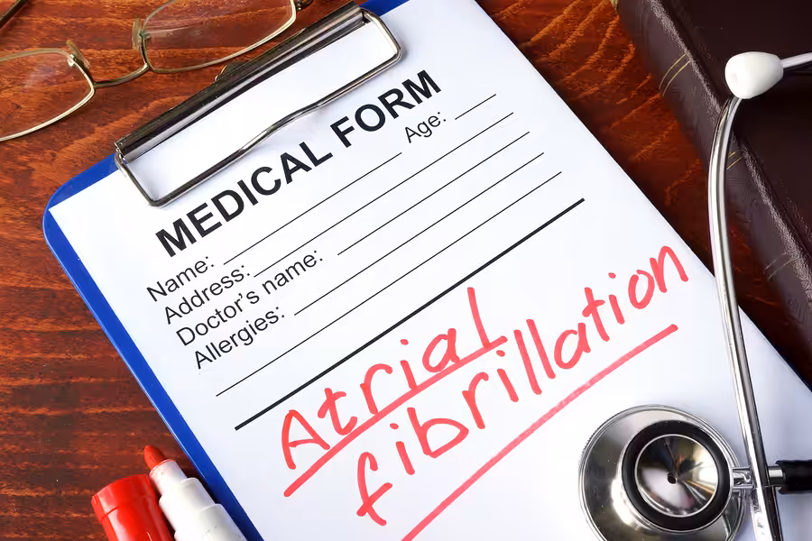 A medical form on a clipboard with Atrial fibrillation written in red marker. A stethoscope, eyeglasses, and a brown book are nearby on a wooden desk. The form fields include name, age, doctor’s name, and allergies.