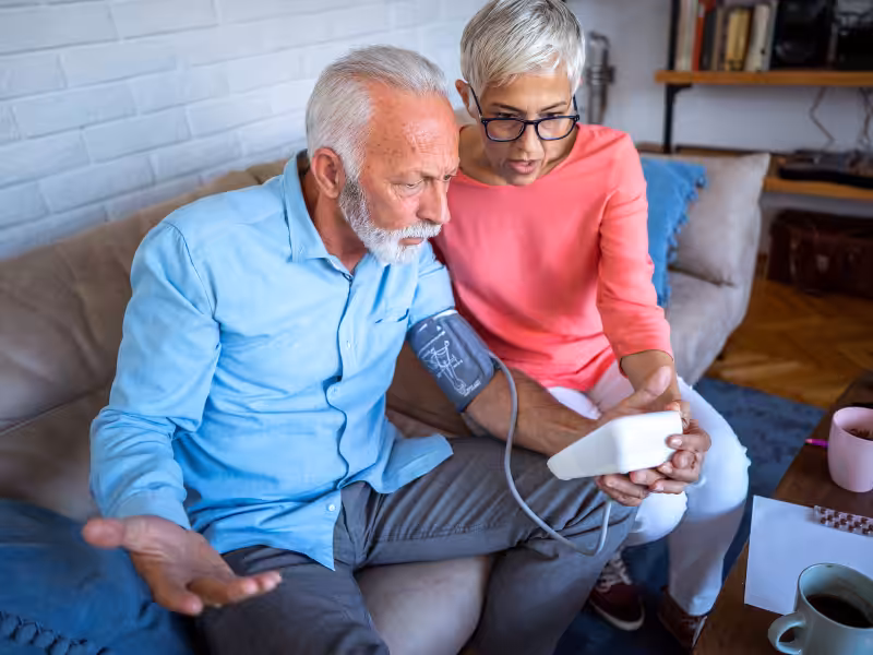 An older man sits on a couch with a blood pressure cuff on his arm, looking confused, while a woman next to him examines the blood pressure monitor, both appearing concerned.