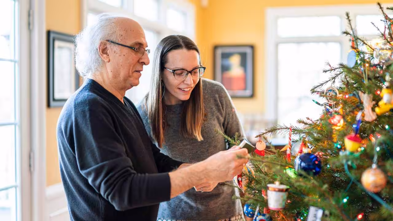 An older man and a young woman decorate a Christmas tree together in a brightly lit room, smiling as they hang ornaments.