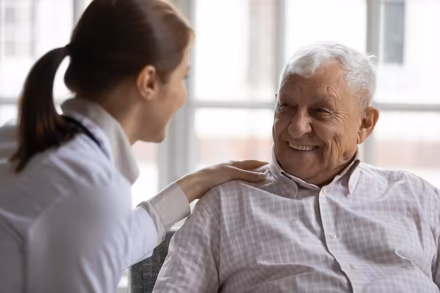 A young female caregiver smiles and rests her hand on the shoulder of an elderly man, who is sitting and smiling back at her in a bright, cozy room.