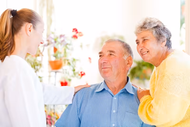 A smiling elderly couple talks with a young female caregiver, who is gently resting her hand on the mans shoulder. They appear to be in a bright, cheerful indoor setting with plants in the background.