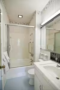A small bathroom with a walk-in shower with glass doors, a white toilet, a white vanity with a sink, a wall mirror, and towels hanging on a rack. The walls have light-colored wallpaper with vertical stripes.