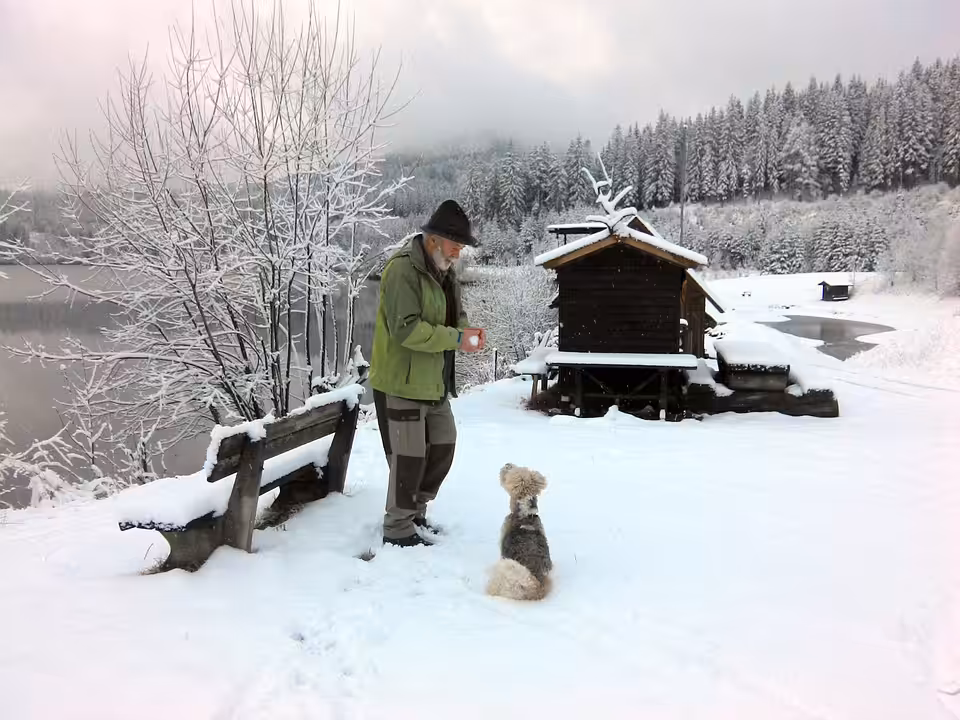 An older man in winter clothes holds a mug while standing in snow next to a small dog. They are near a wooden cabin by a snow-covered lake, surrounded by snowy trees and hills.