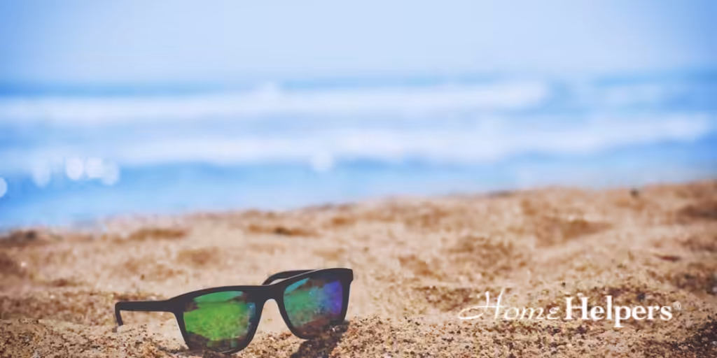 A pair of sunglasses lies on sandy beach with the ocean and blue sky blurred in the background. The words Home Helpers are written in white in the bottom right corner.