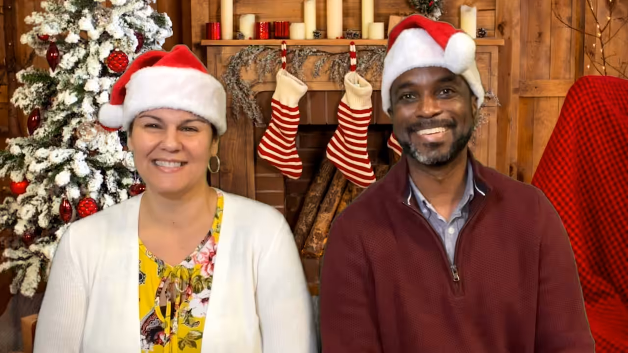 Two people wearing Santa hats smile in front of a festive fireplace decorated with stockings, candles, and a Christmas tree adorned with ornaments and lights.