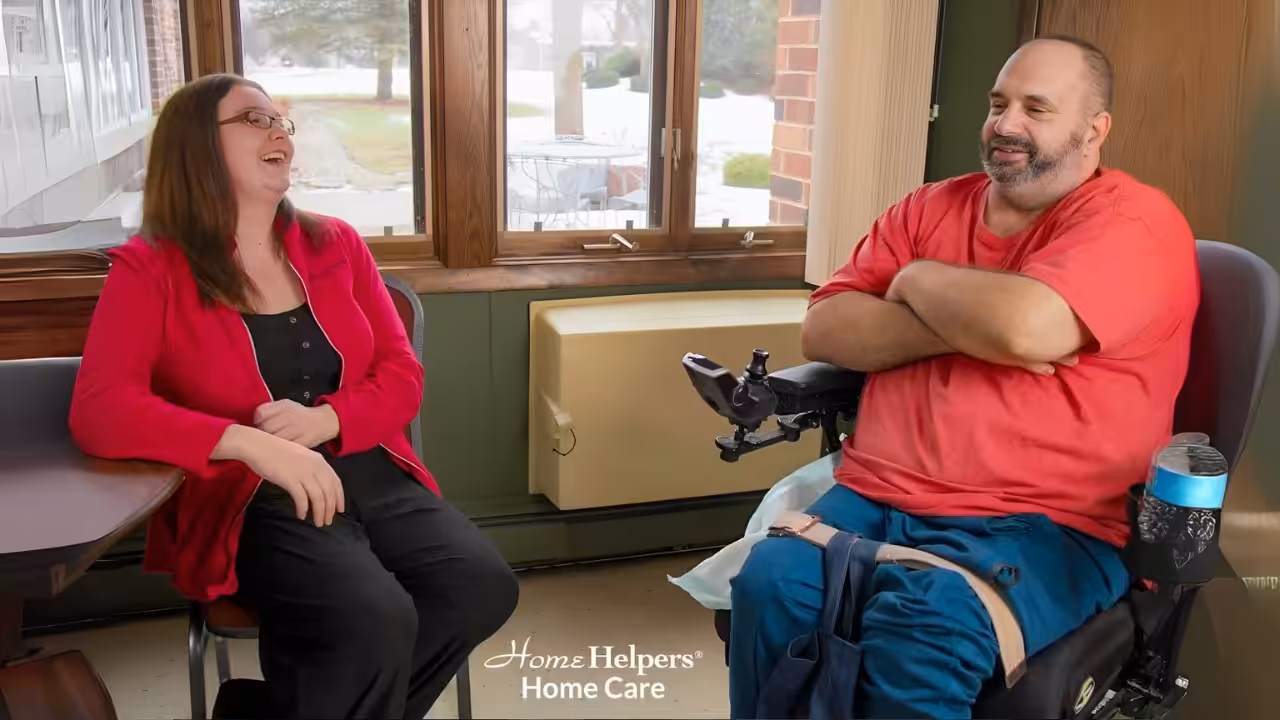 A woman and a man in a wheelchair are smiling and laughing together indoors by a window. The man is wearing a red shirt and has a drink attached to his wheelchair. The image includes the Home Helpers Home Care logo.