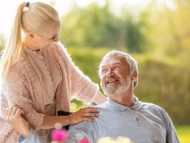 A smiling elderly man sits outside in a wheelchair, looking up at a younger woman who stands beside him with her hand on his shoulder. They are both smiling and surrounded by greenery.