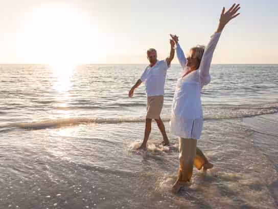 An older couple walks hand in hand along the shoreline at sunset, smiling and raising their arms joyfully as waves gently lap at their feet.
