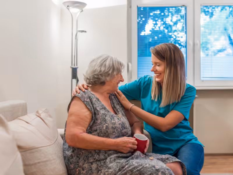 A smiling caregiver in blue scrubs sits next to an elderly woman holding a red mug on a couch, gently touching her shoulder. They are inside a cozy, well-lit room with large windows in the background.